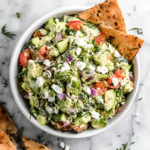 Avocado feta dip with tomatoes, red onion, herbs, and pita chips in a white bowl
