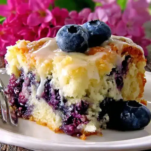 A slice of blueberry boyfriend bait cake with fresh blueberries and lemon glaze on a white plate