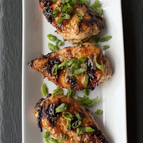 Juicy chicken breasts marinated and broiled, garnished with green onions on a white plate
