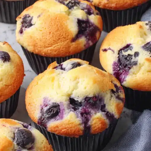 Freshly baked Greek yogurt blueberry muffins with juicy berries, golden tops, and black liners on marble