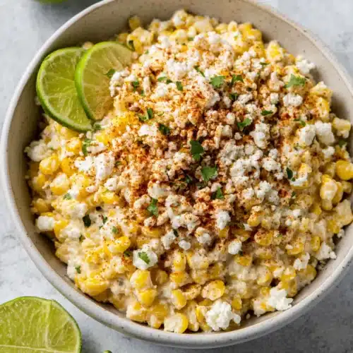 Homemade street corn dip with lime wedges and cotija cheese in a white bowl