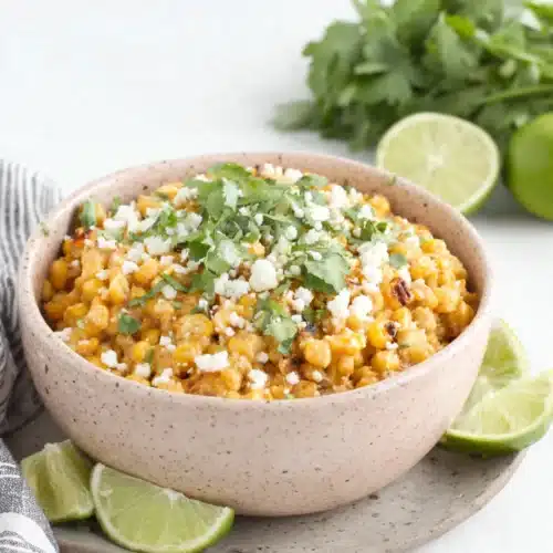 Mexican street corn casserole in a pink bowl with lime wedges and cilantro