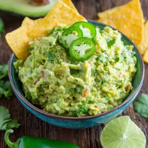 Spicy guacamole in a white bowl with jalapeño slices and tortilla chip