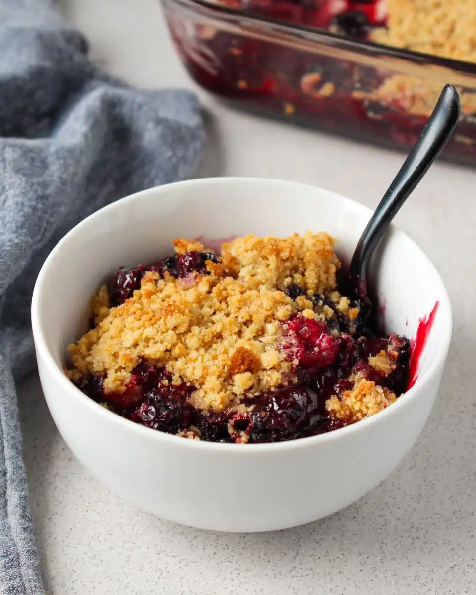 Triple berry crisp in a white bowl with a black spoon, golden crumble topping and juicy berries.