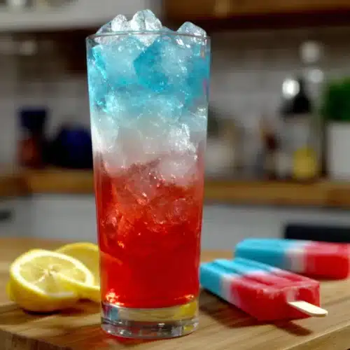 Bomb Pop drink with red, white, and blue layers, lemon slices, and popsicles on a wooden board.