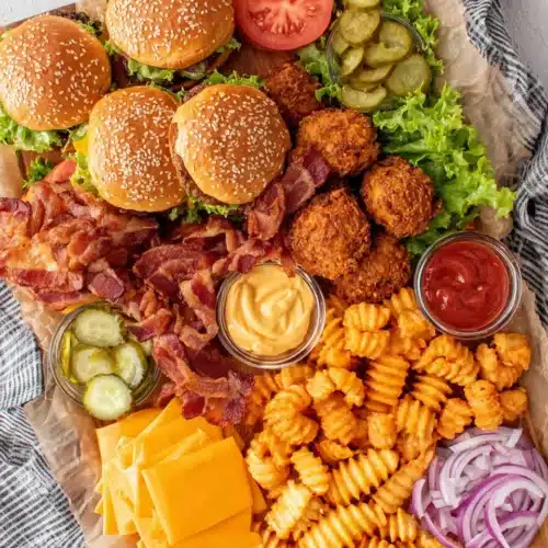 Burger board with patties, buns, cheese, bacon, veggies, condiments, and fries