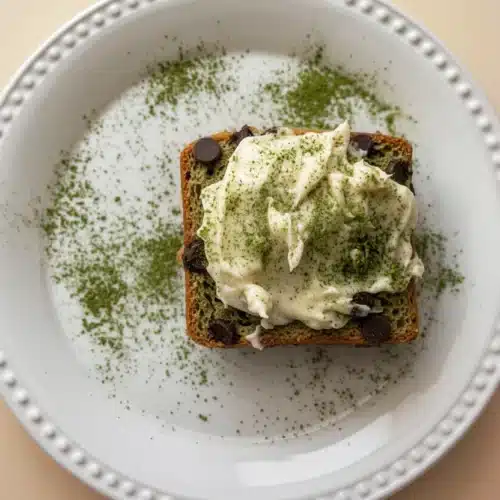 Chocolate chip matcha bread slice with white frosting and matcha powder on a white plate.