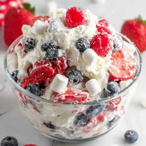 Red white and blue cheesecake salad with strawberries, blueberries, and marshmallows in a glass bowl