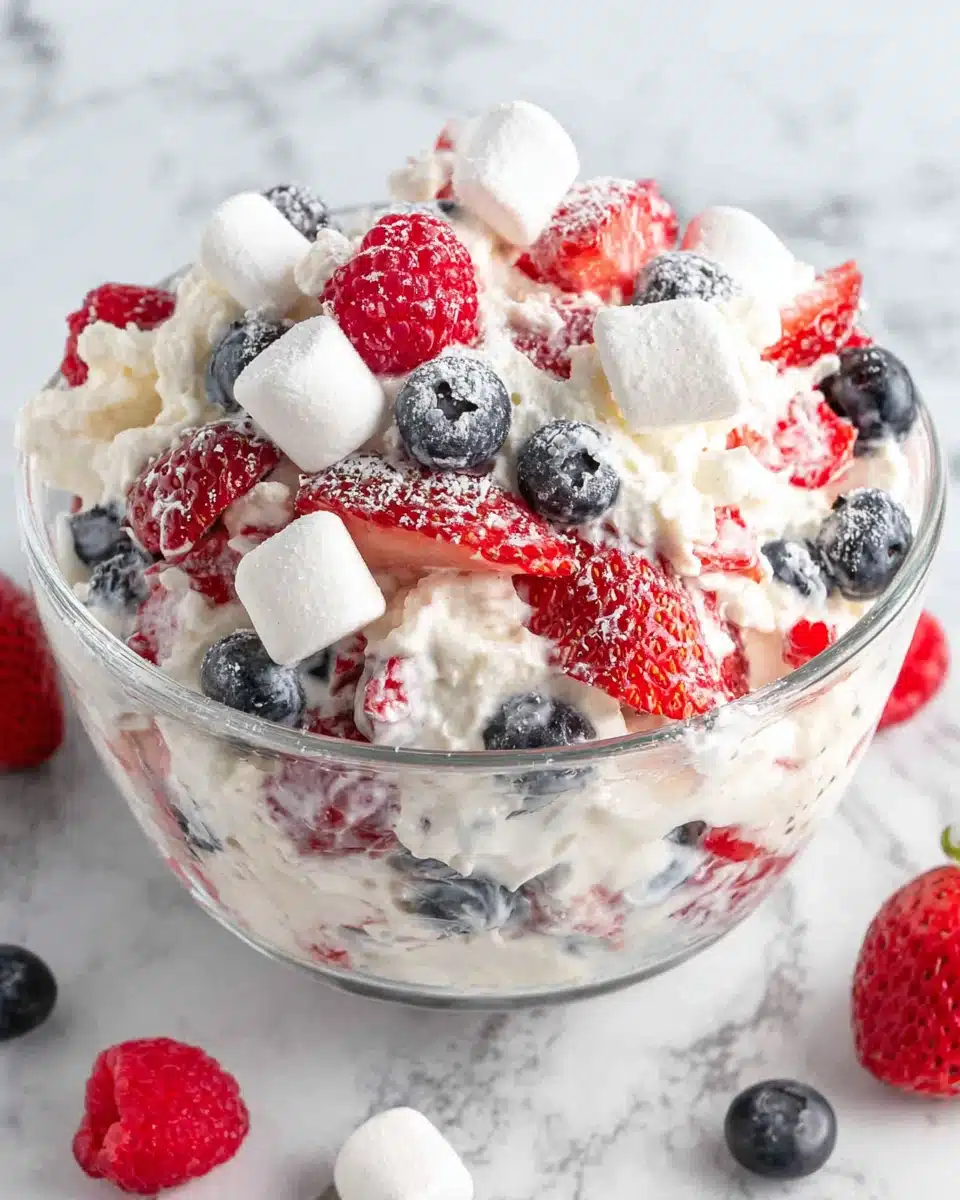 Red white and blue cheesecake salad with strawberries, blueberries, marshmallows, and cream in a glass bowl